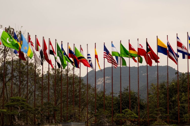 Diversas bandeiras nacionais hasteadas diante de uma paisagem montanhosa, representando cooperação internacional.